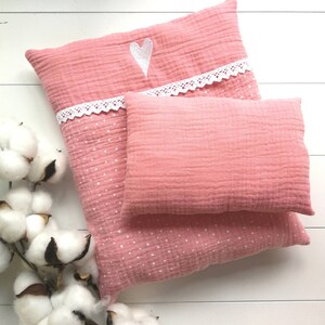 Könnte beinhalten: Zwei rosa Musselin-Kissen, eines rechteckig und eines quadratisch, liegen auf einer weißen Holzoberfläche. Das größere Kissen hat eine weiße Spitzenborte und ein weißes Herzdesign. Der Stoff hat ein strukturiertes, geknittertes Aussehen mit kleinen weißen Punkten.