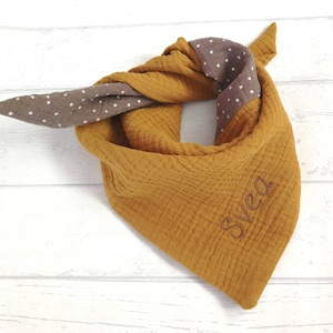 May include: Mustard yellow and brown bandana-style scarf. The scarf is made of a textured fabric and features a brown section with white polka dots. The name "Svea" is embroidered in brown thread on the yellow section.