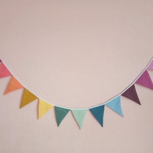 Puede incluir: Una guirnalda de tela colorida con banderas triangulares en tonos de rojo, naranja, amarillo, verde, azul, morado y rosa. Las banderas están enhebradas en una cuerda blanca y están decoradas con lunares blancos.