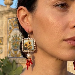 May include: Gold earrings featuring a square sunflower print, framed by small pearls. A large white pearl and red coral elements hang below. The earrings are worn by a person.