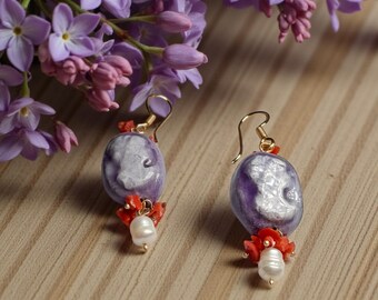 May include: A pair of handcrafted earrings featuring oval, lavender-colored beads with a white design. The earrings have gold-colored hooks, red coral accents, and a dangling white pearl. Lilac flowers are in the background.