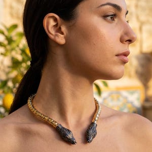 May include: A gold and silver snake necklace with two snake head terminals. The necklace is worn around the neck and has a textured, scale-like pattern. The snake heads are detailed with dark accents.