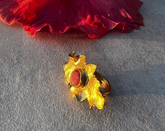 May include: A gold ring with a pink gemstone set in a flower-shaped design. The ring is on a gray textured surface.
