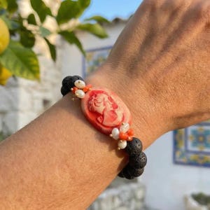 Könnte beinhalten: Armband mit roter Kamee mit weißem Profil, umgeben von weißen Perlen und orangefarbenen Perlen. Das Armband ist auf schwarzen Lavaperlen aufgefädelt, wodurch ein Kontrast von Farben und Texturen entsteht. Einzigartiger Schmuck.