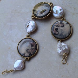 Puede incluir: Una pulsera de estilo vintage con colgantes redondos dorados con imágenes de querubines, intercalados con grandes perlas blancas irregulares. La pulsera tiene una estética antigua delicada, con un cierre de gancho.