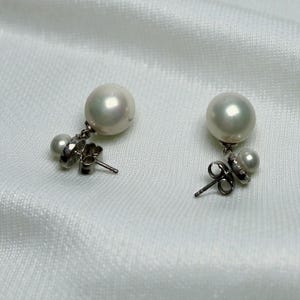 May include: A pair of pearl earrings with a post back. Each earring features a large, round, white pearl suspended below a smaller pearl and a silver-toned metal setting. The earrings are displayed on a white, textured surface.
