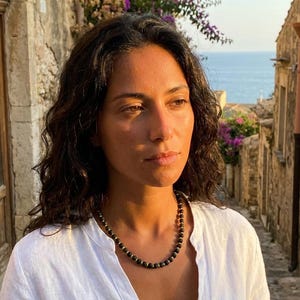 Può includere: Una collana di perline nere con dettagli dorati. La collana è indossata sopra una camicia di lino bianca. L'immagine è scattata all'aperto con un edificio in pietra e il mare sullo sfondo. La collana è un gioiello.