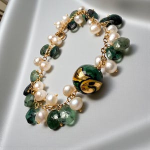 Könnte beinhalten: Ein goldfarbenes Armband mit grünen Edelsteinen, weißen Perlen und einer großen, runden, grünen Perle mit goldenen Akzenten. Das Armband-Design umfasst eine filigrane Kette, die einen Kontrast aus Texturen und Farben erzeugt. Ein Schmuckstück.
