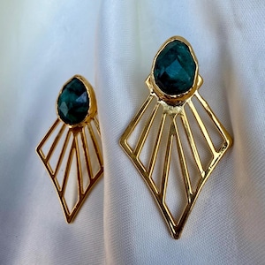 Può includere: Un paio di orecchini color oro con un design geometrico. Ogni orecchino presenta una pietra preziosa verde scuro a forma di goccia incastonata in una lunetta dorata. Gli orecchini hanno una cornice a forma di diamante con linee radiali.