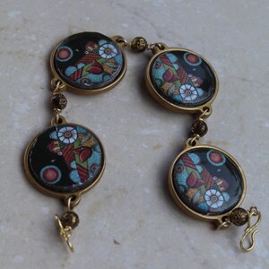 Puede incluir: Un brazalete vintage con cuatro colgantes circulares esmaltados. Cada colgante presenta un diseño floral y figurativo en rojo, azul y verde sobre un fondo negro. Los colgantes están unidos por eslabones dorados y un cierre.