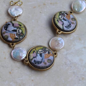 Puede incluir: Un collar dorado con colgantes circulares con pinturas en miniatura y perlas blancas redondas e iridiscentes. Las pinturas representan figuras con ropa colorida. El collar se exhibe sobre una superficie clara y texturizada.