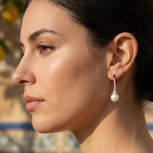 May include: Elegant silver earrings with a dangling white pearl and sparkling accents. The earrings are shown on a person's ear, with a blurred background of lemons and a decorative tile wall.
