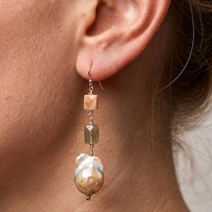 May include: Gold-toned drop earrings featuring a combination of natural elements. The earrings showcase a light peach-colored square bead, a gray-green square bead, and a large, irregular-shaped pearl. The design is suspended from a gold hook.