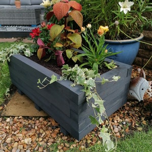 May include: A dark grey wooden planter box filled with a variety of plants, including ivy, red and orange flowers, and green foliage. The planter is sitting on a bed of small stones.
