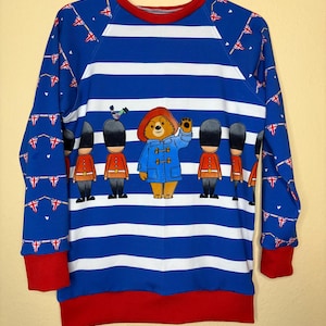Könnte beinhalten: Blau-weiß gestreiftes Sweatshirt mit einem winkenden Paddington Bär-Motiv, umgeben von Spielzeugsoldaten. Die Ärmel und der Halsausschnitt sind rot eingefasst, und die Ärmel haben ein Muster aus Union Jack-Flaggen.