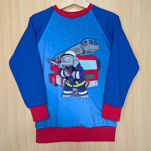 Könnte beinhalten: Blaues Langarmshirt mit rotem Ausschnitt, Bündchen und Saum. Es zeigt einen Cartoon-Feuerwehr-Elefanten und ein Feuerwehrauto. Die Ärmel sind einfarbig blau.