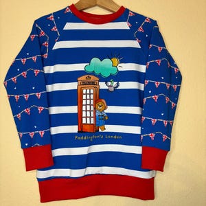 Könnte beinhalten: Blau-weiß gestreiftes Sweatshirt mit roten Bündchen und Kragen. Es zeigt eine Paddington Bär Grafik vor einer roten Telefonzelle, mit Sonne, Wolke und Taube. Die Ärmel haben ein Muster aus Union Jack Flaggen.