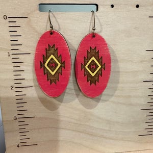May include: A pair of red oval earrings with a geometric design in brown and yellow. The earrings are hanging on a wooden display board with a ruler and the text "by JCLA".