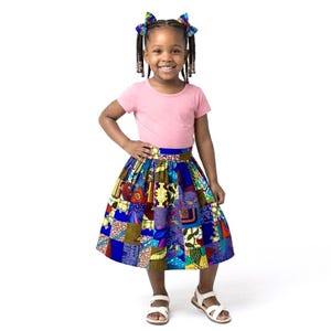 May include: A young girl wearing a pink t-shirt and a colorful patchwork skirt. The skirt features a vibrant mix of blue, yellow, red, and purple patterns. She is also wearing white sandals and has her hair styled with colorful bows.