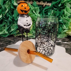 Op de afbeelding: Een glazen beker met een zwart spinnenwebontwerp, een bamboe deksel met een gat voor een rietje en een bijpassend oranje rietje. Op de achtergrond staat een keramiek pompoenfiguur met een zwarte hoed en een wit shirt.