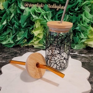 Op de afbeelding: Een glazen pot met een zwart-wit spinnenwebontwerp, een bamboe deksel en een roestvrijstalen rietje. De pot staat op een wit marmeren oppervlak met een bruin bamboe deksel en een helder glazen rietje.