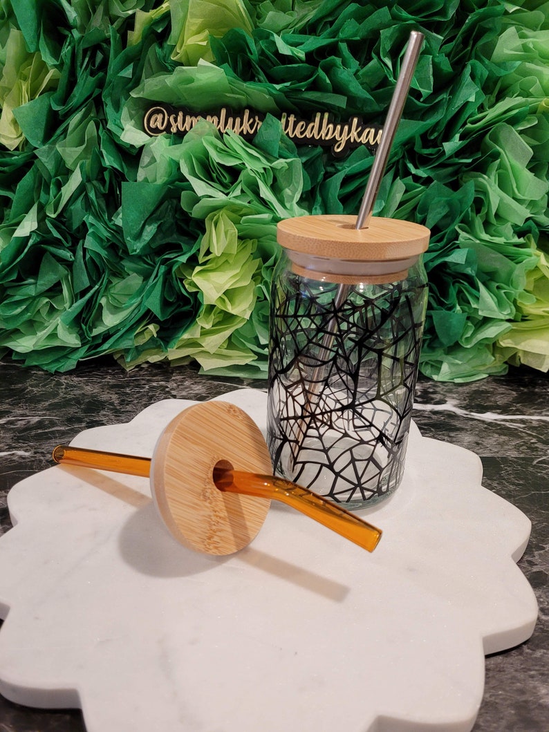 Op de afbeelding: Een glazen pot met een zwart-wit spinnenwebontwerp, een bamboe deksel en een roestvrijstalen rietje. De pot staat op een wit marmeren oppervlak met een bruin bamboe deksel en een helder glazen rietje.