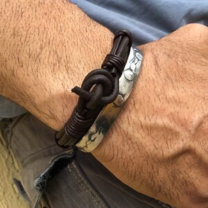 May include: A brown leather bracelet with a silver metal band. The leather is braided and has a knot at the end. The metal band is textured and has a hammered finish.