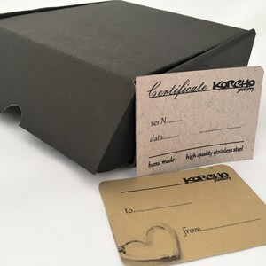 May include: A brown cardboard box with a certificate of authenticity for a piece of jewelry. The certificate is labeled "Certificate KORCHO Jewelry" and includes the text "ser.N...", "data...", "hand made", "high quality stainless steel", and "KORCHO Jewelry". A second card is labeled "to..." and "from..." with a heart design.