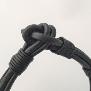 May include: A black leather bracelet with a knotted design. The bracelet is made of multiple strands of leather that are woven together.