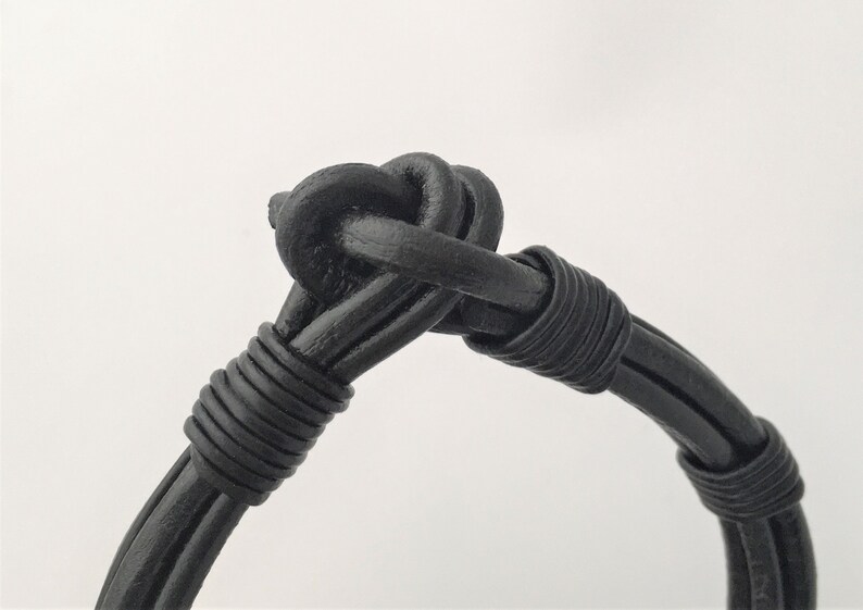 May include: A black leather bracelet with a knotted design. The bracelet is made of multiple strands of leather that are woven together.