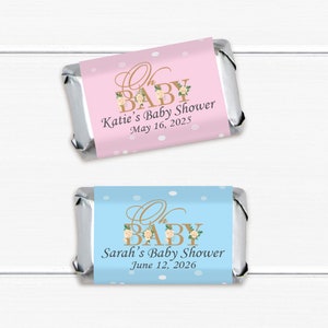 May include: Two pink and blue baby shower favor labels with gold lettering and floral accents. The text on the pink label reads "Oh Baby Katie's Baby Shower May 16, 2025". The text on the blue label reads "Oh Baby Sarah's Baby Shower June 12, 2026".