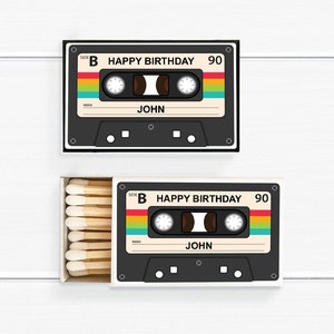 May include: Two matchboxes with a retro cassette tape design. The cassette tapes are black with a colorful stripe and the text "HAPPY BIRTHDAY 90" and "JOHN". One box is open, revealing wooden matches.