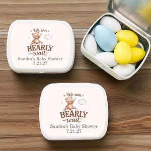 Könnte beinhalten: Weiße, rechteckige Babyparty-Gastgeschenk-Dosen mit dem Text "We can bearly wait" und einer Teddybär-Grafik. Eine Dose ist geöffnet und zeigt bunte Süßigkeiten. Das Datum der Babyparty ist auf der Dose aufgedruckt.