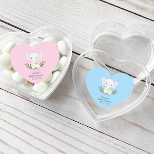 Puede incluir: Dos recipientes de plástico transparentes en forma de corazón con tapas. Cada recipiente tiene una pegatina con un lindo elefante de dibujos animados, flores y el texto "Nicole's Baby Shower 07.21.24". Un recipiente tiene una pegatina rosa y el otro tiene una pegatina azul.