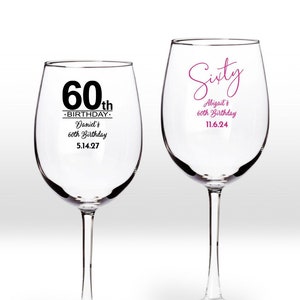 May include: Two clear wine glasses with personalized 60th birthday messages. The glass on the left says "60th Birthday - Daniel's 60th Birthday 5.14.27". The glass on the right says "Sixty - Abigail's 60th Birthday 11.6.24".
