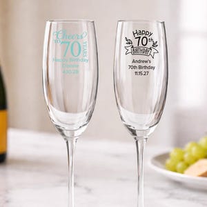 May include: Two clear champagne flutes with personalized text for a 70th birthday. One flute has "Cheers to 70 Years" and the name "Dianne" with a date. The other has "Happy 70th Birthday" and the name "Andrew" with a date. A bottle of champagne is in the background.
