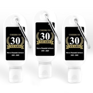 Könnte beinhalten: Drei weiße Handdesinfektionsmittel-Flaschen mit silbernen Karabinerhaken. Jede Flasche hat ein schwarzes Etikett mit goldenem Text "CHEERS TO 30 YEARS" und "Morse Financial Advisors 1995-2025".