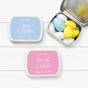 May include: Three small, white metal tins with rounded corners. Two tins are closed, one blue and one pink, each with custom text. The third tin is open, revealing colorful candies. The text on the blue tin reads "Mint to Be Carrie & Matthew March 10, 2027."