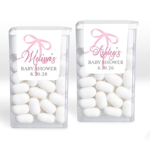 May include: Two clear plastic containers filled with white mints. Each container has a white label with a pink bow and text that reads "Melissa's BABY SHOWER 8.20.28" and "Ashley's BABY SHOWER 6.10.26".