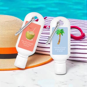 May include: Two white travel-sized bottles with silver carabiner clips. Each bottle has a label with text and a tropical illustration. One label is coral with a coconut drink, the other is light blue with a palm tree.