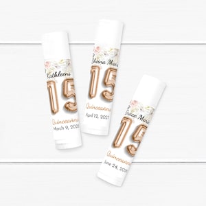May include: Three white lip balm tubes with personalized labels for a quinceañera. Each label features a floral design with gold balloon numbers "15" and the name of the quinceañera, the date of the event, and the words "Quinceañera".