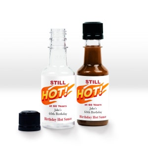 May include: Two clear glass bottles with black caps and labels. The labels are white with red and orange text that reads "STILL HOT!" and "at 60 Years Jake's 60th Birthday Birthday Hot Sauce".