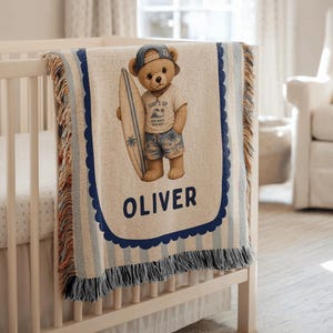 May include: A woven throw blanket featuring a tan teddy bear wearing a blue baseball cap, a t-shirt with a wave graphic, and patterned shorts while holding a surfboard. The name OLIVER is printed in large dark blue letters below the bear.