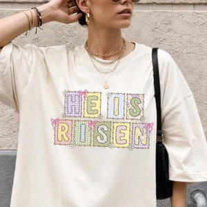 Puede incluir: Camiseta de gran tamaño de color marfil con el texto "HE IS RISEN" en colores pastel. La camiseta se combina con pantalones cortos a cuadros rojos y blancos. La modelo lleva gafas de sol y joyas.
