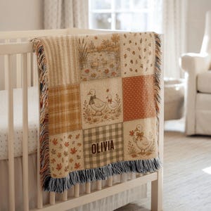 Puede incluir: Una manta tejida colocada sobre una cuna blanca, con un diseño de patchwork con motivos otoñales, que incluye patos, hojas caídas y cuadrados a cuadros. La manta incluye el nombre OLIVIA impreso en una fuente de color marrón oscuro.