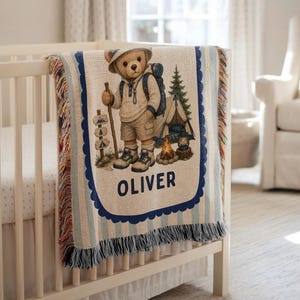Puede incluir: Una manta tejida que muestra un oso de peluche con equipo de senderismo, mochila, bastón, tienda de campaña y fogata. El diseño incluye una señal con iconos de árboles y el nombre OLIVER en letras azul oscuro, con bordes de flecos decorativos.