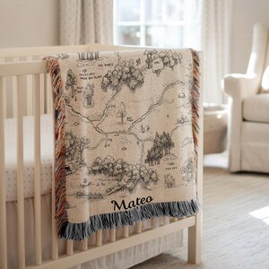 Puede incluir: Una manta de bebé beige y negra con un mapa temático y el nombre "Mateo" impreso, colocada sobre una cuna blanca. La manta presenta un diseño detallado de bosque y río con bordes con flecos en varios colores. La cuna está en una habitación luminosa con una ventana.