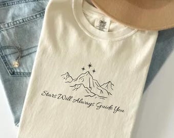 Op de afbeelding: Een crèmekleurig t-shirt met een zwarte lijntekening van bergtoppen en sterren, met de tekst "Stars Will Always Guide You" eronder gedrukt. Het t-shirt wordt gecombineerd met een lichtblauwe spijkerbroek en een beige vilten hoed.