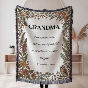 Puede incluir: Una manta de color crema con la palabra "GRANDMA" en letras negras grandes. La manta presenta ilustraciones florales y botánicas en tonos marrones, verdes y melocotón. El texto dice: "Ella habla con sabiduría, y la instrucción fiel está en su lengua. - Proverbios 31:26."