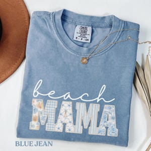May include: A blue jean Comfort Colors t-shirt with the words "beach MAMA" in white script and patchwork letters. The letters are decorated with seashells, gingham, and stripes. A gold necklace with a pendant is draped across the shirt.